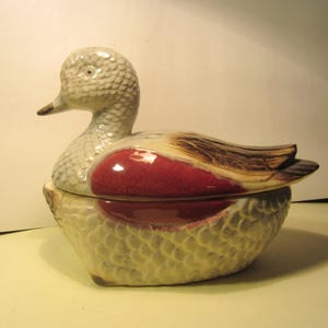 May include: A ceramic duck-shaped container with a lid. The duck has a gray head and a red and brown body. The container is light yellow with a textured, scale-like pattern.