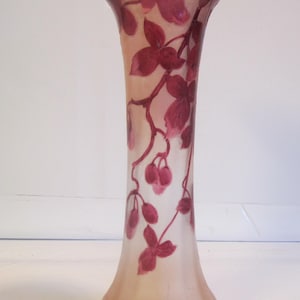 May include: A tall, elegant glass vase with a flared top and a rounded base. The vase is adorned with a delicate floral design in shades of burgundy and pink, featuring leaves and berries. The vase has a frosted appearance.