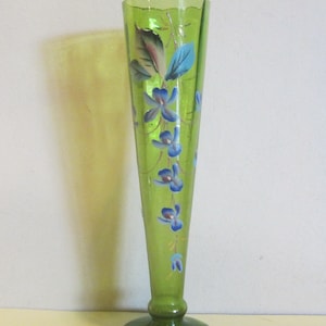 Pode incluir: Um vaso de vidro verde com uma base alargada e um pescoço longo e fino. O vaso é decorado com flores pintadas à mão em azul e verde.