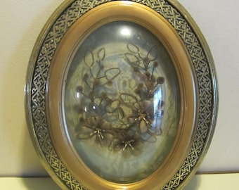 Puede incluir: Un marco de imagen ovalado con un marco dorado y un patrón decorativo. El marco tiene una cubierta de vidrio y contiene un arreglo de flores secas.