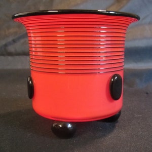 Puede incluir: Un jarrón de vidrio rojo con rayas negras y puntos negros. El jarrón tiene tres patas negras.