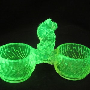 May include: A glowing green glass double salt cellar with a decorative bird-like figure in the center. The two bowls have a textured design. The object is set against a black background.