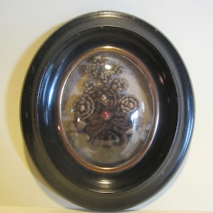 May include: A black oval frame with a glass cover encloses a cluster of brown hair woven into a flower shape with white beads. The hair art is displayed on a white background.