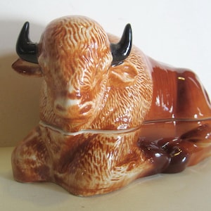 May include: A ceramic bison butter dish with a brown and white glaze. The bison is lying down with its head resting on its front legs. The dish is divided into two sections, one for the butter and one for the lid.