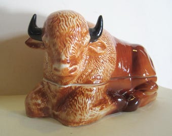 May include: A ceramic bison butter dish with a brown and white glaze. The bison is lying down with its head resting on its front legs. The dish is divided into two sections, one for the butter and one for the lid.