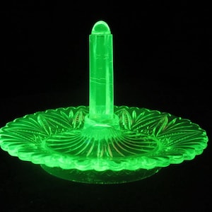 May include: A glowing green glass ring holder with a central post and a decorative, scalloped base. The base features a detailed, etched pattern. The ring holder is illuminated against a black background.