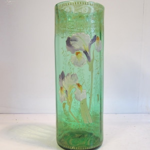 Könnte beinhalten: Eine zylindrische Vase aus grünem Glas mit einem bemalten Blumenmuster aus weißen und violetten Schwertlilien mit gelben Zentren. Das Glas ist durchscheinend und der obere sowie untere Rand sind mit einem dezenten geometrischen Muster verziert.