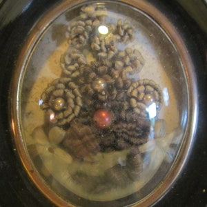 May include: A framed oval-shaped piece of art featuring a cluster of brown hair woven into flower shapes with pearl and red bead accents.