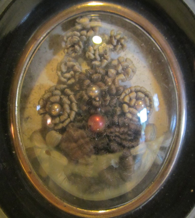 May include: A framed oval-shaped piece of art featuring a cluster of brown hair woven into flower shapes with pearl and red bead accents.