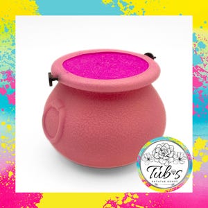 May include: A pink cauldron-shaped bath bomb container with a textured surface and a bright pink interior. The container has a small handle and a decorative emblem on the side. The container is labeled "Tub's Bath Bombs".