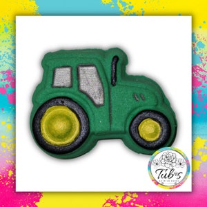 Puede incluir: Una bomba de baño verde con forma de tractor con ruedas y detalles amarillos y negros. El tractor tiene una ventana plateada y una chimenea negra. La bomba de baño está sobre un fondo blanco con salpicaduras de pintura coloridas.