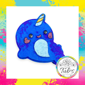 Könnte beinhalten: Eine narwalförmige Badebombe in leuchtendem Blau mit glitzerndem Finish. Sie hat ein goldgestreiftes Horn, einen hellblauen Bauch, schwarze Augen und rosa Wangen. Ein kreisförmiges Logo zeigt florale Linienkunst und den Text Tub's Bathtub Bombs.