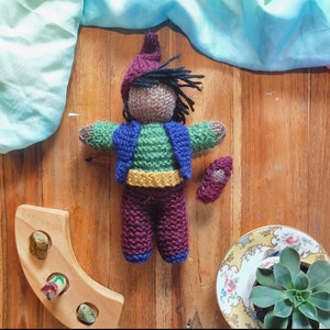 May include: A knitted doll with brown skin, green and blue clothing, and a red hat. The doll is sitting on a wooden surface.