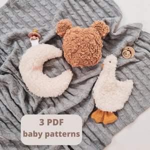 Könnte beinhalten: Drei Plüschtiere für Babys: ein brauner Bär, ein weißer Halbmond und ein weißes Küken. Die Spielzeuge liegen auf einer grauen Strickdecke. Der Text "3 PDF Baby Muster" befindet sich auf einem weißen Rechteck.