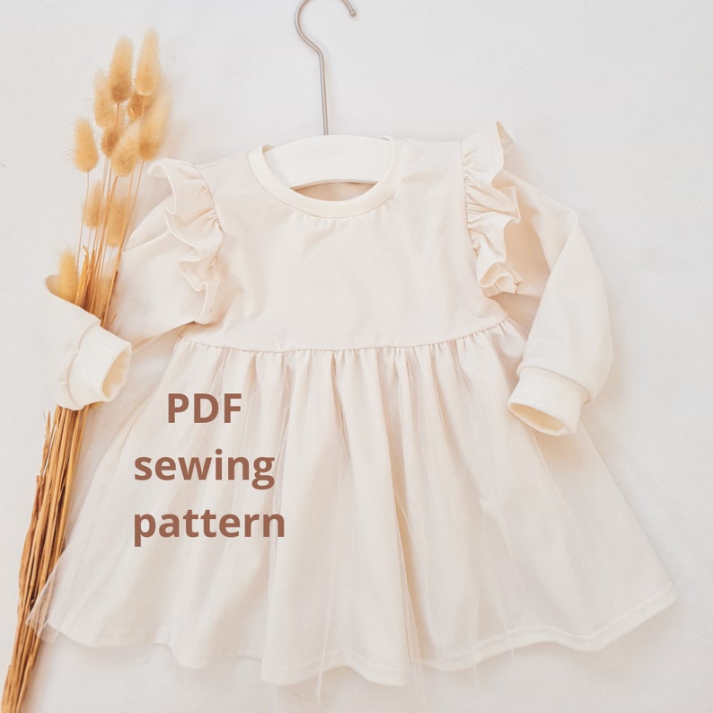May include: Cream-colored baby dress with long sleeves and a tulle skirt, hanging on a white hanger. The dress features ruffle detailing on the shoulders. Text on the dress reads "PDF sewing pattern".