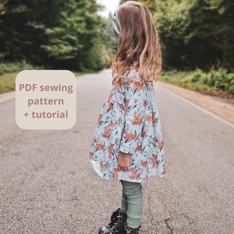 May include: A young person stands on a road, wearing a light blue dress with a fox and floral pattern. The dress has long sleeves and a ruffled hem. The text "PDF sewing pattern + tutorial" is in a light beige square. The person is also wearing green leggings and black boots.
