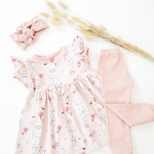 May include: A baby girl's outfit set featuring a pink dress with a bunny print, matching leggings, and a headband. The dress has ruffled sleeves and a gathered skirt. The leggings are ribbed, and the headband has a bow.