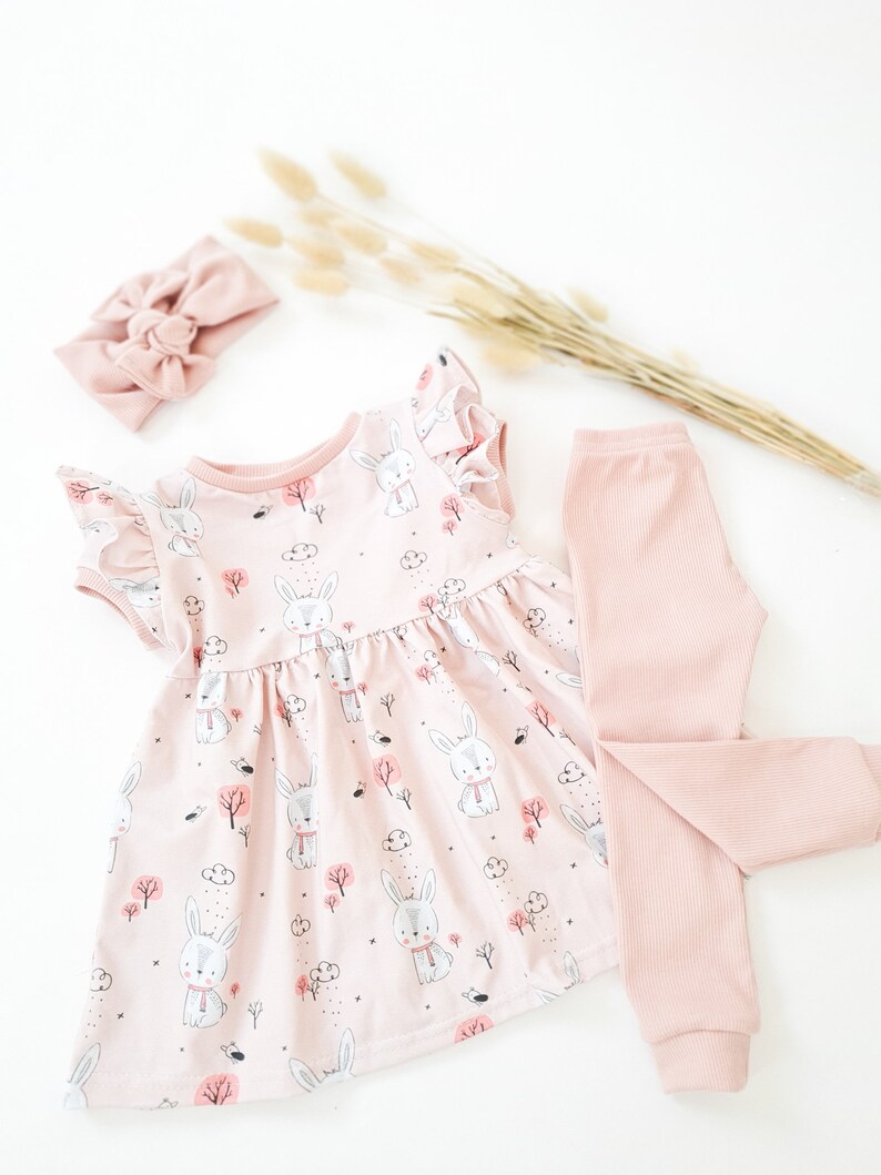 May include: A baby girl's outfit set featuring a pink dress with a bunny print, matching leggings, and a headband. The dress has ruffled sleeves and a gathered skirt. The leggings are ribbed, and the headband has a bow.