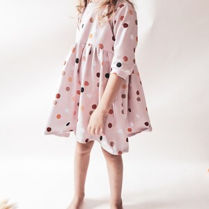 May include: A light pink dress with a round neckline and three-quarter sleeves. The dress is adorned with a pattern of colorful polka dots in shades of brown, orange, and black. The dress has a flared skirt and is knee-length.