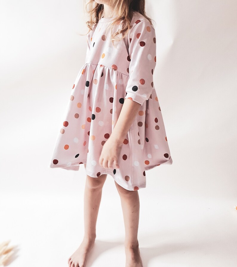 May include: A light pink dress with a round neckline and three-quarter sleeves. The dress is adorned with a pattern of colorful polka dots in shades of brown, orange, and black. The dress has a flared skirt and is knee-length.
