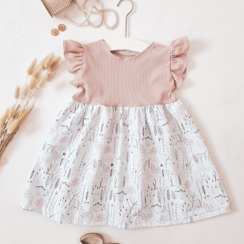 May include: A light pink baby dress with a floral print skirt and ruffled sleeves. The bodice is a solid light pink ribbed knit fabric. The skirt features a white background with a delicate pattern of gray and pink botanical elements.