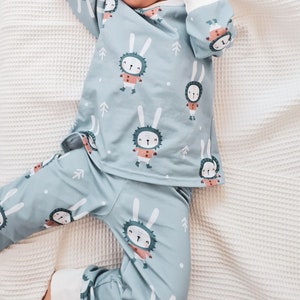 May include: A light blue baby onesie with a white trim and a repeating pattern of white rabbits wearing red scarves and boots. The rabbits are surrounded by small white dots and green pine trees.