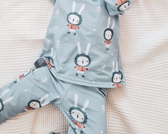 May include: A light blue baby onesie with a white trim and a repeating pattern of white rabbits wearing red scarves and boots. The rabbits are surrounded by small white dots and green pine trees.