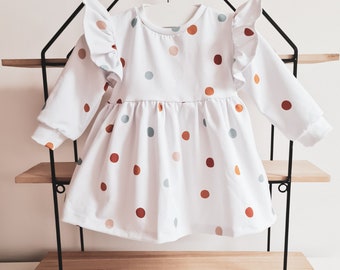 May include: White long-sleeved dress with a polka dot pattern in shades of orange, brown, and blue. The dress features ruffled sleeves and a gathered skirt. The dress is displayed on a black metal shelf.
