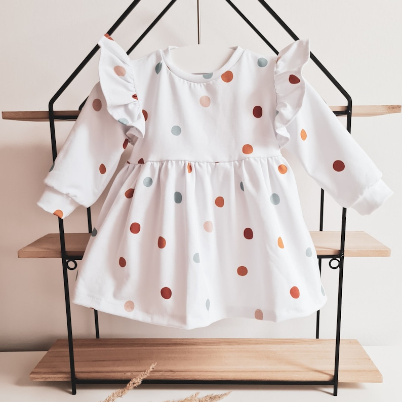 May include: White long-sleeved dress with a polka dot pattern in shades of orange, brown, and blue. The dress features ruffled sleeves and a gathered skirt. The dress is displayed on a black metal shelf.