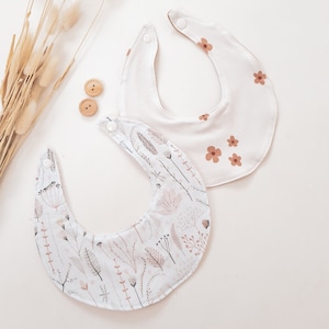 Puede incluir: Dos baberos de bebé blancos con patrones florales. Un babero tiene un patrón floral marrón y el otro tiene un patrón floral marrón claro.
