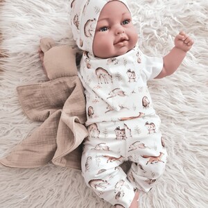 May include: A baby doll wearing a white onesie with a brown and white woodland animal print. The onesie has long sleeves and pants. The doll is also wearing a brown and white hat with a woodland animal print. The doll is lying on a white fluffy surface.
