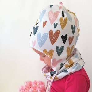 Peut inclure: Un bonnet blanc à capuche avec un motif de cœurs colorés. Le bonnet a un cordon de serrage rose et est porté par un enfant.