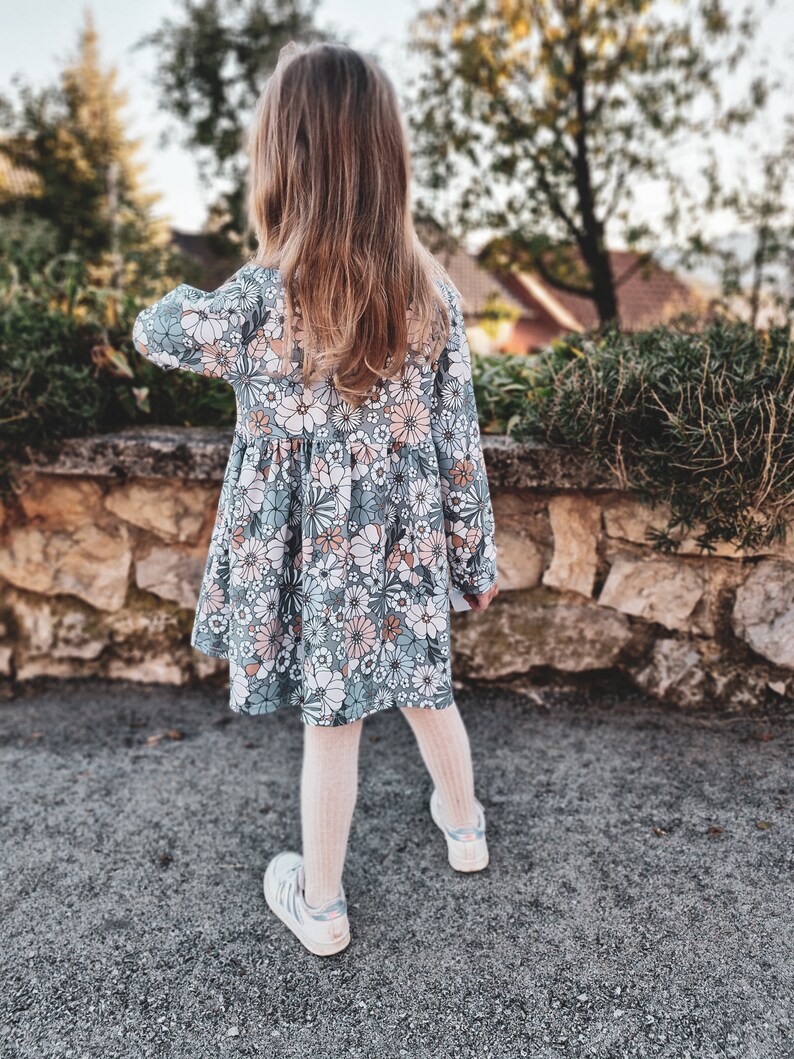 May include: A young child wearing a long-sleeved dress with a floral pattern in shades of blue, pink, and white. The child is also wearing white sneakers and light-colored tights. The dress has a round neckline and a gathered skirt.