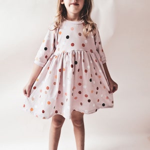 May include: A light pink dress with a round neckline and three-quarter sleeves. The dress features a gathered waist and is adorned with a pattern of various-sized polka dots in shades of brown, orange, and white. The dress is designed for children.