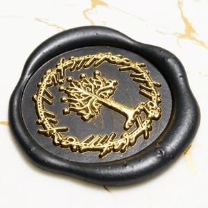 May include: A circular black wax seal featuring a raised gold-coloured design of a tree centred within a ring inscribed with Elvish script. The wax has an irregular, organic edge and a smooth, matte finish, highlighting the metallic gold details.