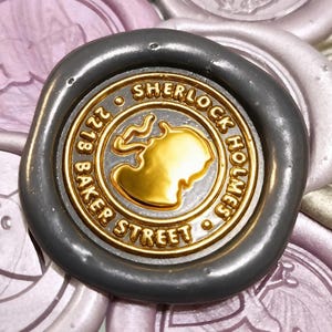 May include: A dark grey wax seal featuring a gold-coloured centre with a silhouette of a person smoking a pipe. The text reads Sherlock Holmes 221B Baker Street. It is surrounded by several other pale pink and metallic wax seals.