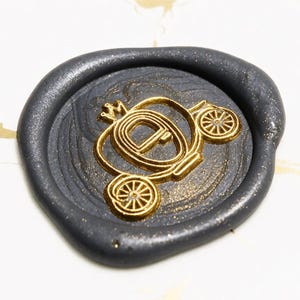 May include: A round wax seal in a dark charcoal grey colour with a subtle glittery finish. The centre features a raised, metallic gold embossed design of a classic horse-drawn carriage with detailed wheels and a curved roof structure.