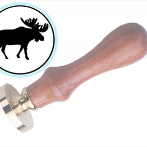 May include: A wax seal stamp with a wooden handle and a brass seal. The seal features a black silhouette of a moose inside a white circle with a light blue border. The handle is a light brown color.