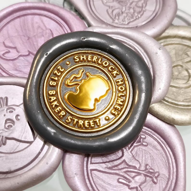 May include: A dark grey wax seal featuring a gold-coloured centre with a silhouette of a person smoking a pipe. The text reads Sherlock Holmes 221B Baker Street. It is surrounded by several other pale pink and metallic wax seals.