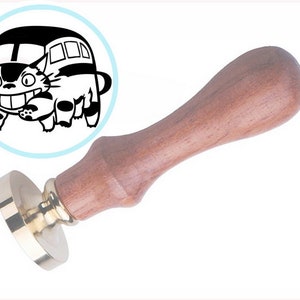 May include: A wax seal stamp featuring a wooden handle and a brass head. The stamp design is a circular black and white graphic of a cat-shaped bus with multiple legs, large eyes, and a wide grin, set against a light blue circular background.