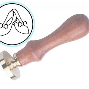 Może przedstawiać: Pieczęć woskowa z drewnianym uchwytem i mosiężną podstawą. Wzór na pieczęci to para butów z kokardkami.