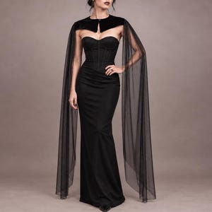 May include: A floor-length black strapless gown featuring a structured corset bodice with vertical seams. The dress is paired with a black velvet bolero-style shoulder wrap that includes long, flowing, sheer black tulle sleeves that extend to the floor.