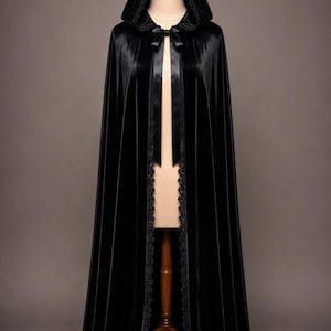 May include: A floor-length black velvet hooded cloak featuring a smooth texture, a pointed hood, and a satin ribbon tie closure at the neck. The front edges and the bottom hem are finished with a delicate black lace trim detail.