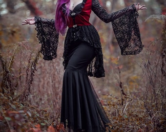 Puede incluir: Una mujer con el pelo largo morado lleva un vestido de encaje negro con un corpiño de terciopelo rojo y un collar de plata. Está de pie en un bosque con follaje otoñal.