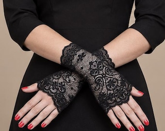 May include: A pair of black fingerless lace gloves featuring a detailed floral pattern and scalloped edges. The gloves cover the wrists and the back of the hands, leaving the fingers and thumbs exposed, and are worn with a black dress.