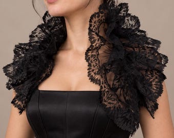 May include: A black lace bolero featuring a high, ruffled collar and intricate floral patterns. The garment is worn over a black, structured, sleeveless bodice with vertical seams, creating a layered look with a distinct, textured lace neckline.