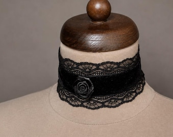 Può includere: Una collana girocollo nera caratterizzata da una fascia centrale in velluto, un delicato bordo in pizzo lungo i margini superiore e inferiore e una piccola decorazione a forma di rosa nera scolpita al centro della sezione in velluto, su un manichino beige.
