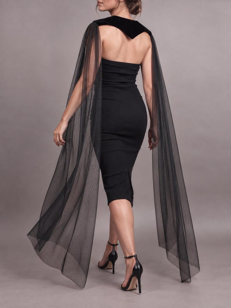May include: A black strapless midi dress featuring a fitted silhouette and a velvet collar detail. Long, sheer black tulle cape panels extend from the shoulders, draping down to the floor. The garment is paired with black high-heeled sandals.