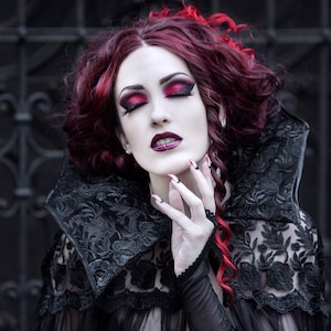 May include: A woman with long, curly red hair wears a black lace dress with a high collar. She has dark eye makeup and red lipstick. Her nails are painted red.
