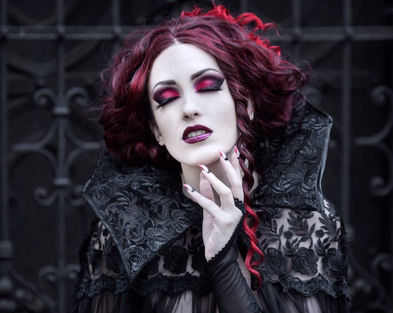 May include: A woman with long, curly red hair wears a black lace dress with a high collar. She has dark eye makeup and red lipstick. Her nails are painted red.